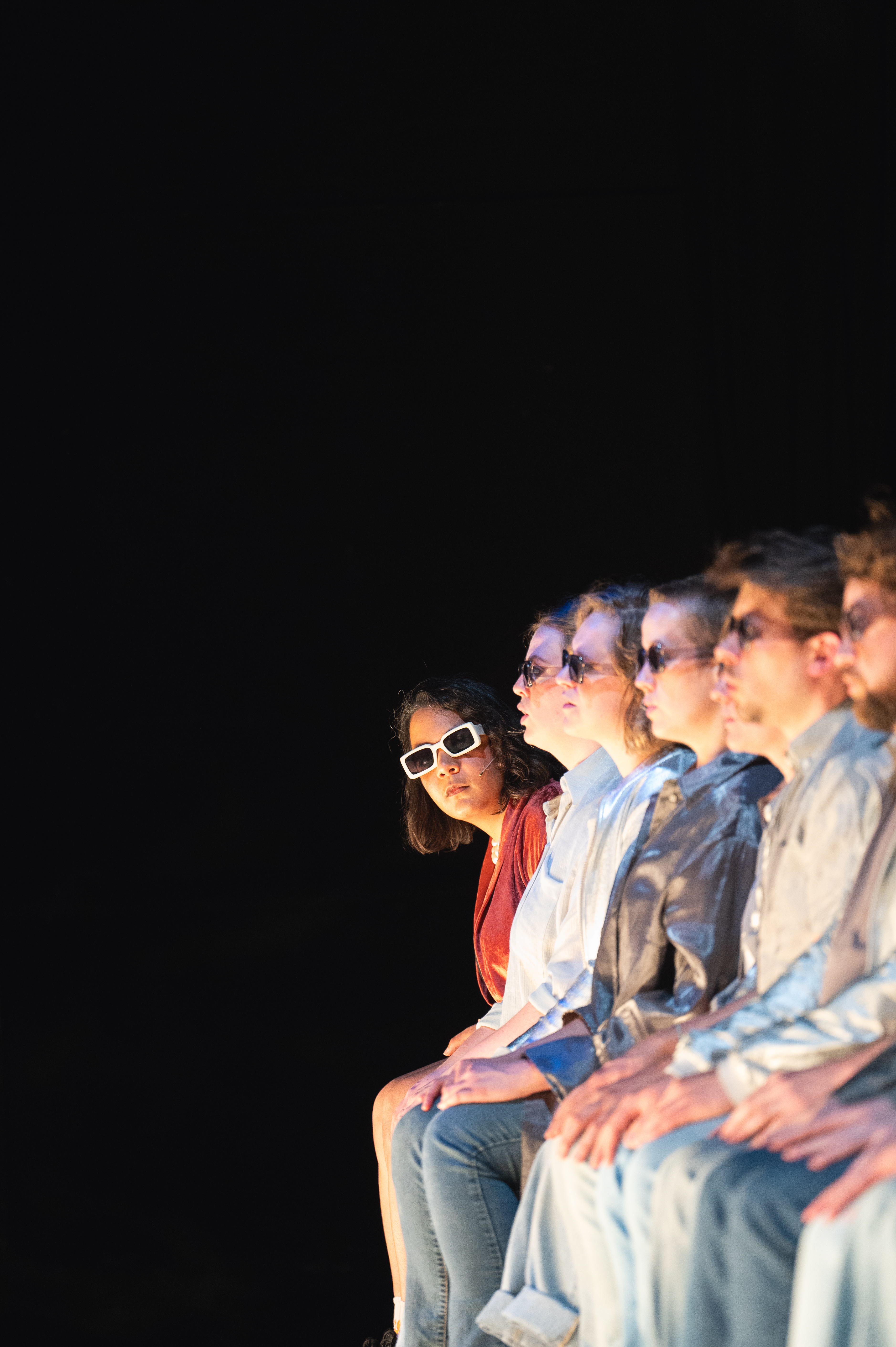 Hinter einer Reihe an singenden Chorsänger:innen im Profil, lugt die Performerin ASJA mit einer weißen Sonnenblille hervor und schaut ins Publikum.