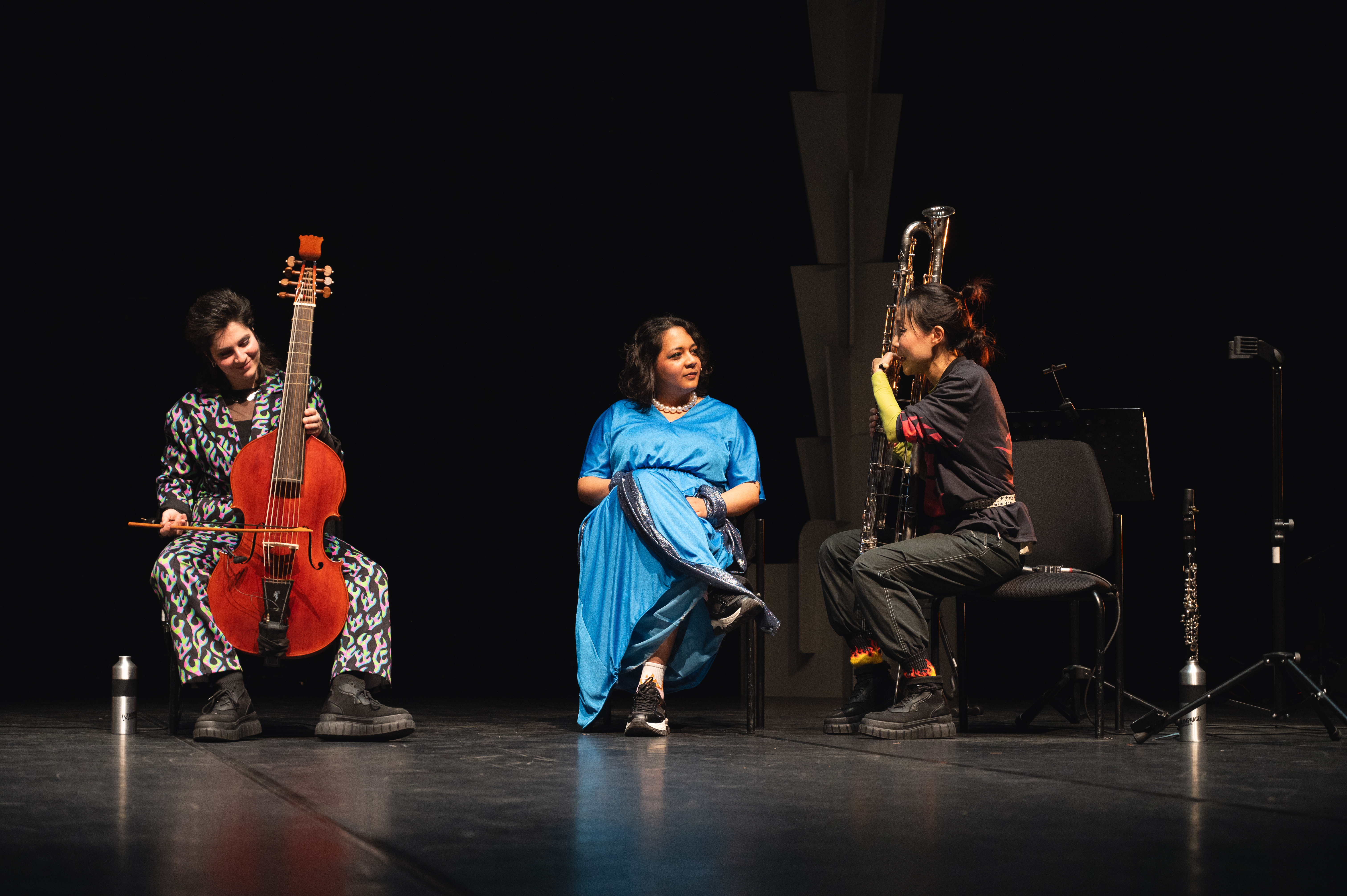 Drei Menschen sitzen auf der Bühne und sind in ein Gespräch vertieft: Die Bassistin Rebecca Lawrence spielt auf einer Gambe und lächelt, ASJA sitzt in einem langen, blauen Kleid in der Mitte und ist  Heni HyunJung Kim zugewandt, die an eine Kontrabassklarinette gelehnt spricht.