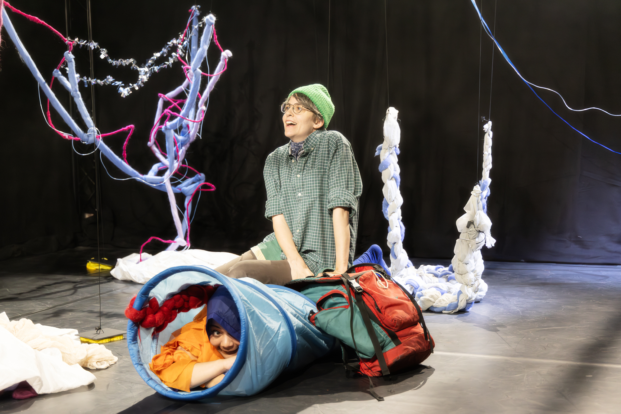 Im Vordergrund sitzt ein Mensch mit Wanderklamotten auf einem Spieltunnel und sieht verzückt aus. In dem Spieltunnel versteckt sich jemand in blau-orangenem Kostüm und grinst. Im Hintergrund sind Bäume aus Stoff, Draht und Wolle.