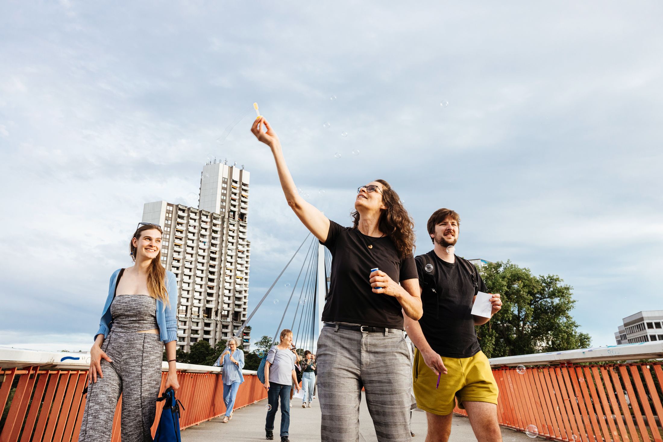 Drei Menschen laufen über eine Brücke, im Hintergrund ist ein Hochhaus. Die Person in der Mitte bläst Seifenblasen.