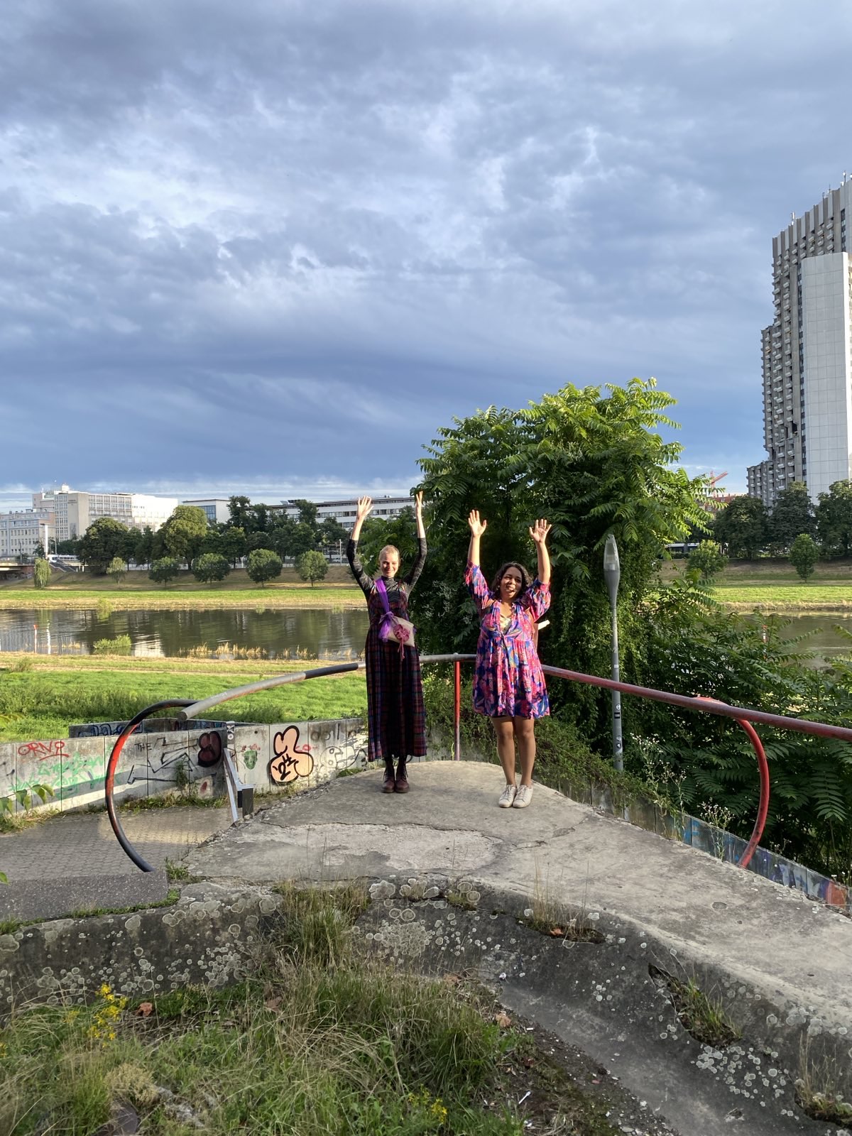 Asja und Lilli stehen vor einem Fluss und heben die Arme.