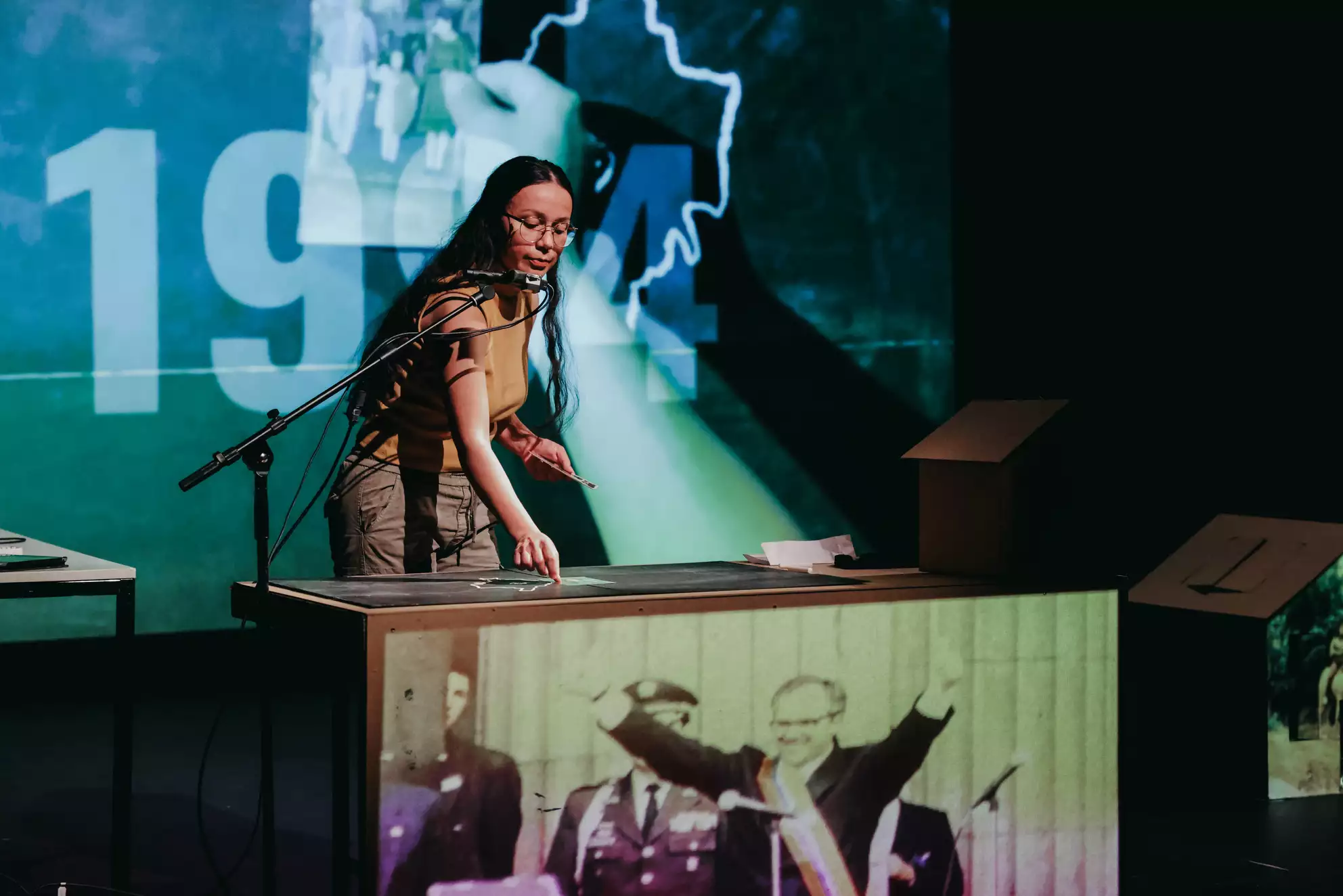 Die Performerin Omayra Martínez Garzón zeichnet mit weißer Kreide auf einem Tisch. Im Hinterdrund ist ein großes Video zu sehen mit der Jahreszahl 1994. Auf die Vorderseite des Tisches ist das Videobild eines Politikers zu sehen.