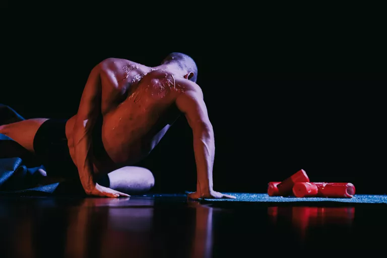 Der Tänzer Mehdi Dahkan sitzt mit dem Rücken zum Publikum am Boden. Auf seinem Rücken sind Kerzenwachsspuren zu sehen. Neben ihm liegen rote, ausgebrannte Kerzen auf grünem Kunstrasen.