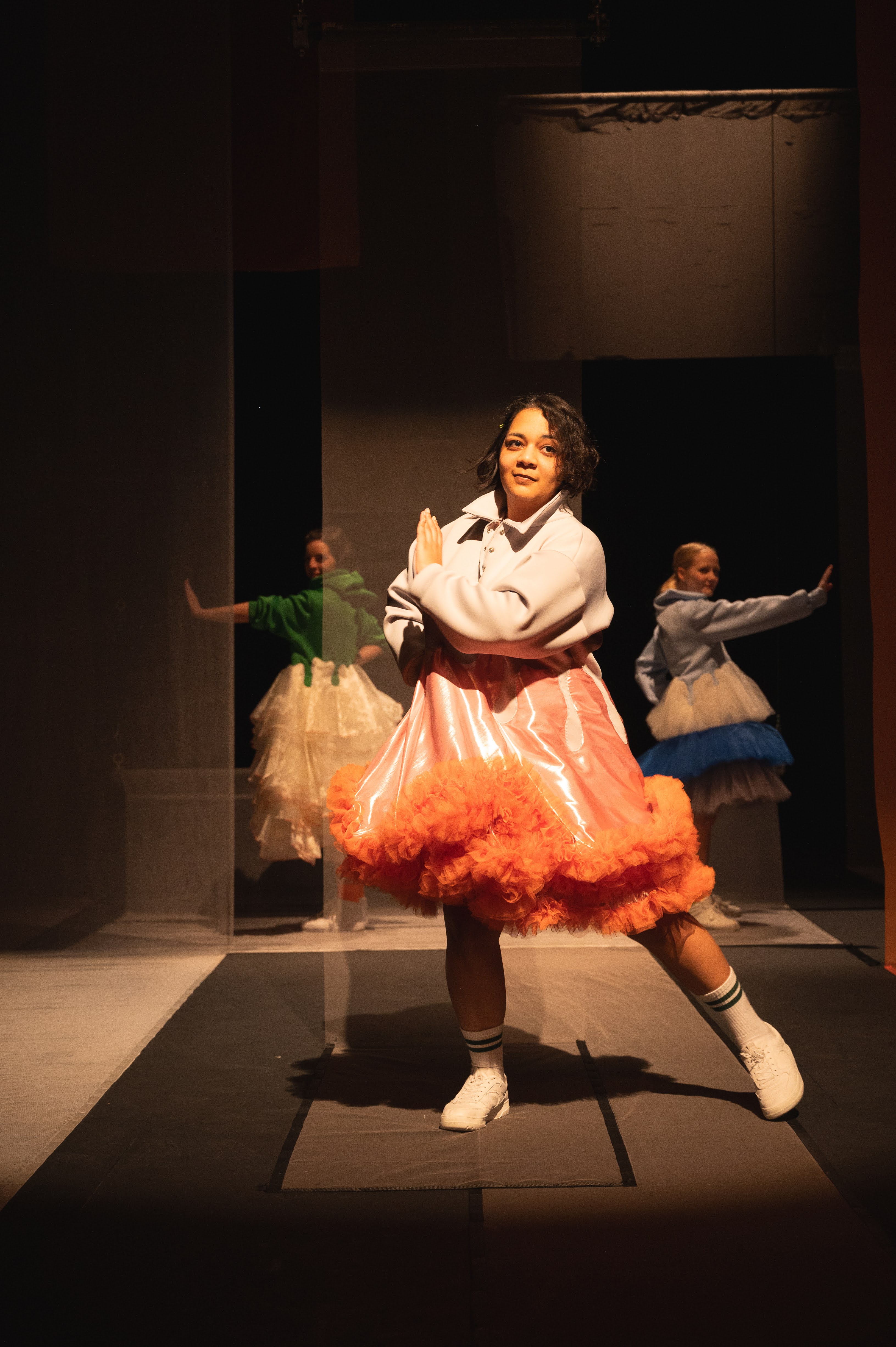 Eine Performerin tanzt im Vordergrund einer Bühne und schaut das Publikum verschmitzt an. Sie trägt ein Kleid mit bauschigem orangenen Tüll, dass sich mit ihr bewegt. Im Hintergrund sind zwei Performerinnen, ebenfalls mit ausgefallenen bunten Kleidern. Sie rahmen mit einer Pose mit ausgestrecktem Arm die Performerin in der Mitte ein.