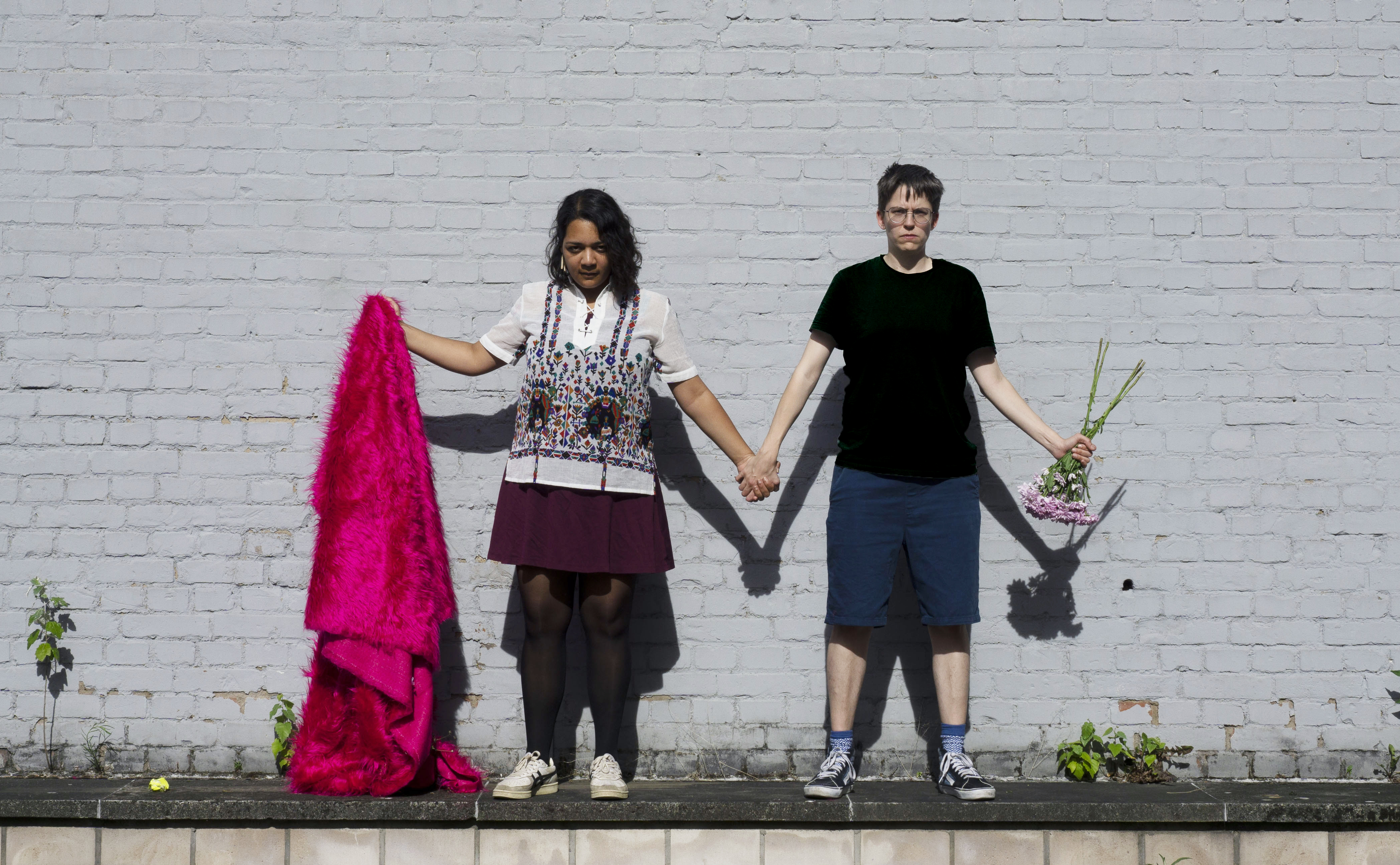 Zwei Menschen stehen vor einer Wand und halten sich an den Händen. Eine Person hält ein pinkes Fell und die andere Person einen umgedrehten Blumenstrauß.